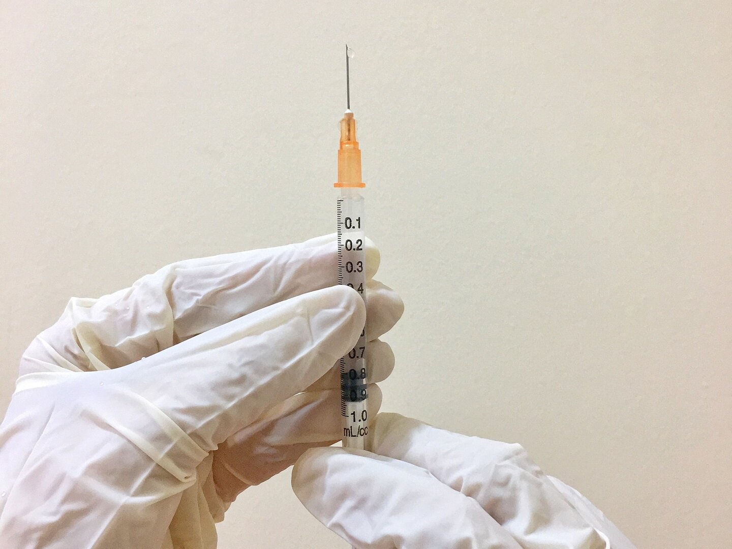 Zdjęcie przedstawia dłonie w rękawiczkach lateksowych trzymające strzykawkę z igłą skierowaną do góry.