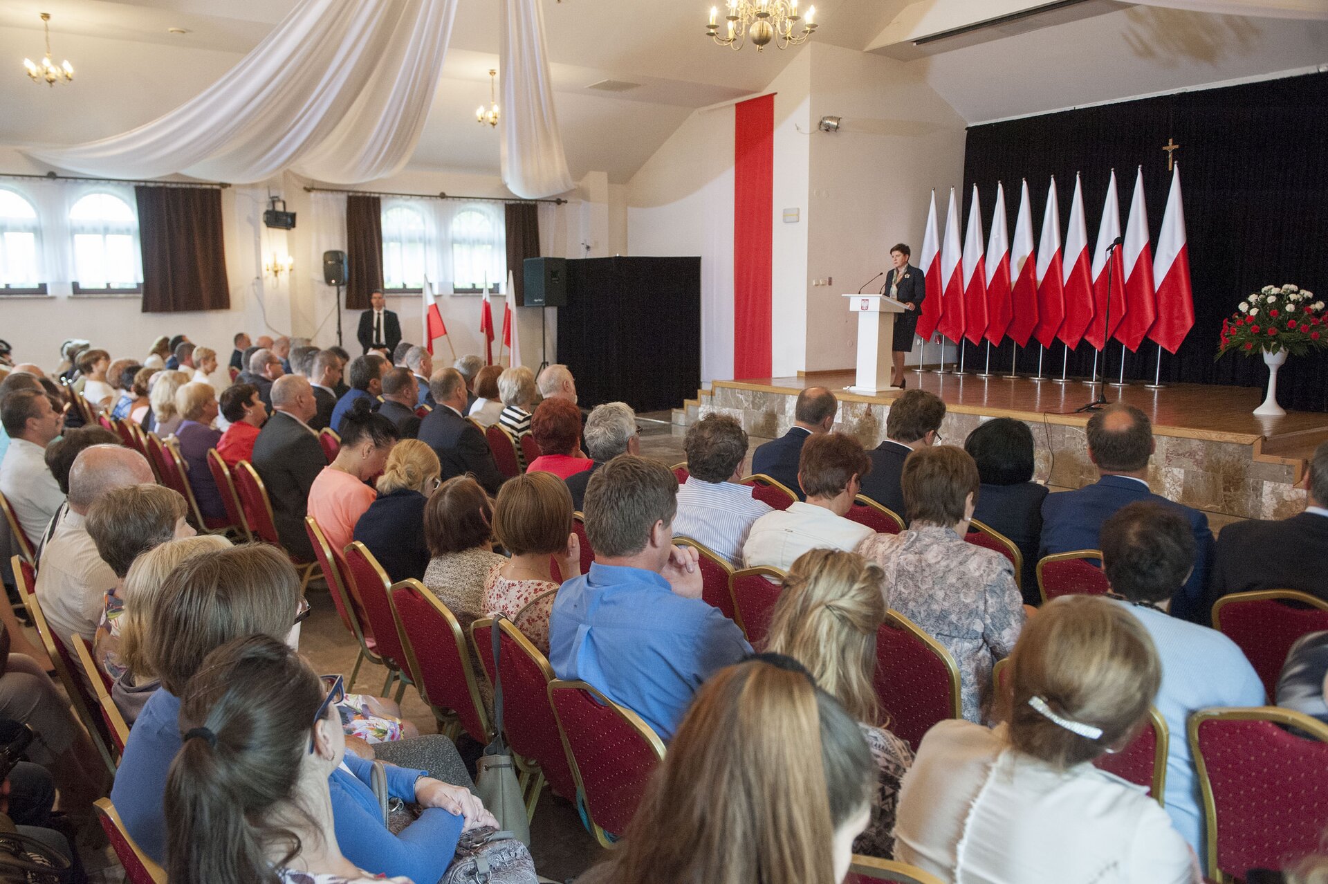 Kolorowa fotografia przedstawiająca przemówienie Beaty Szydło. Na zdjęciu znajduje się sala wypełniona siedzącymi ludźmi. Pani premier przemawia ze sceny, na której ustawiono w rzędzie dziesięć polskich flag oraz umieszczono wazon z czerwono-białymi kwiatami.