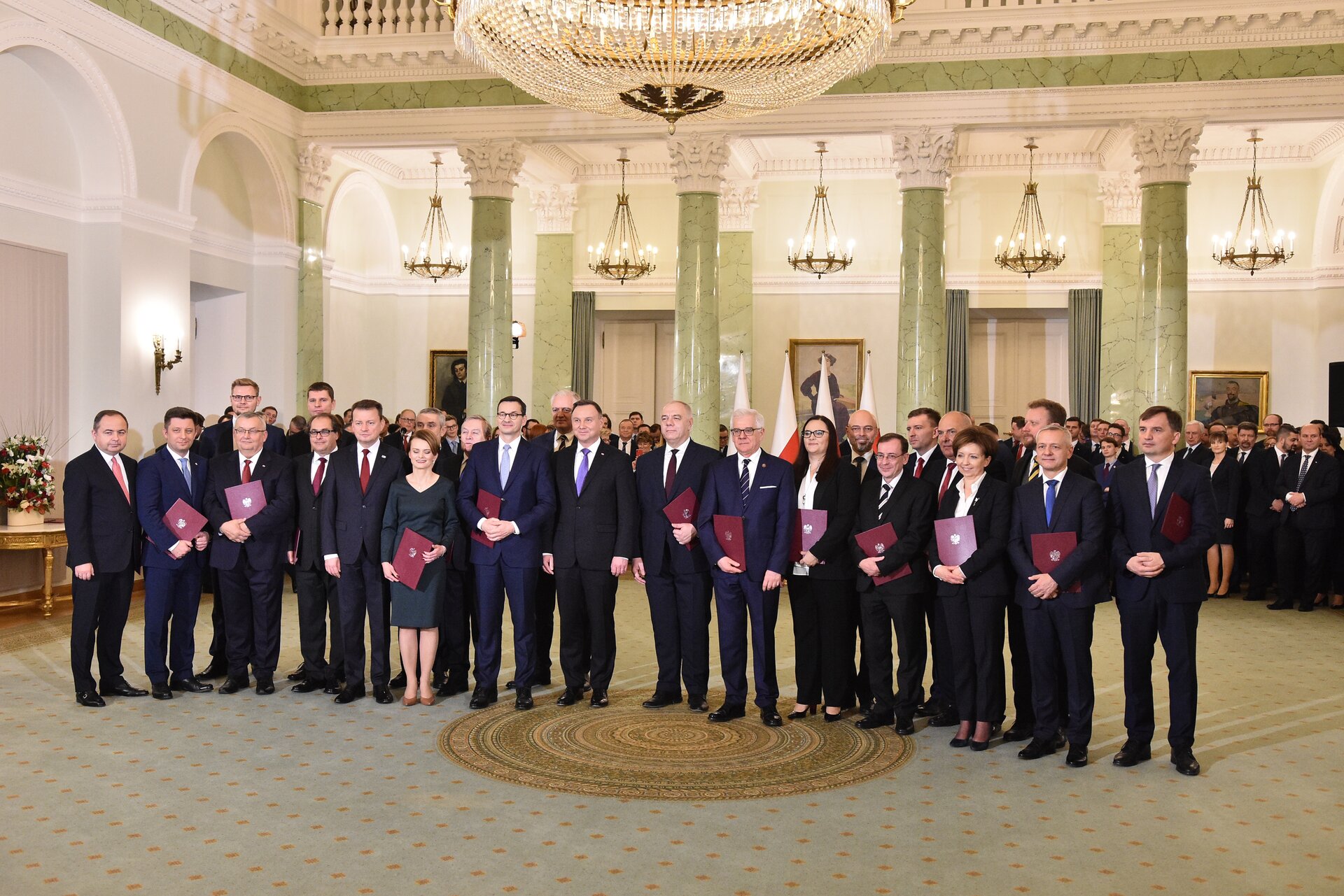 Kliknij żeby powiększyć Na zdjęciu znajdują się: premier Mateusz Morawiecki wraz z Radą Ministrów oraz Prezydentem RP Andrzejem Dudą w Pałacu Prezydenckim. Na pierwszym planie na środku sali stoją obok siebie łącznie 24 osoby, ubrane w eleganckie stroje: mężczyźni w garnitury, kobiety w garsonki. W rękach trzymają teczki z powołaniami na stanowiska. Nad nimi wisi ogromny ozdobny żyrandol. Za nimi widoczne są filary sali, obrazy i pięć ozdobnych wiszących lamp. W tle widoczna jest zgromadzona grupa uczestników uroczystości.
