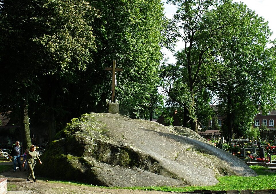 Zdjęcie przedstawia głaz narzutowy. Jeden bok jest wysoki i opada do samego dołu. Na szczycie znajduje się figura krzyż. Wokół znajdują się nagrobki oraz drzewa.