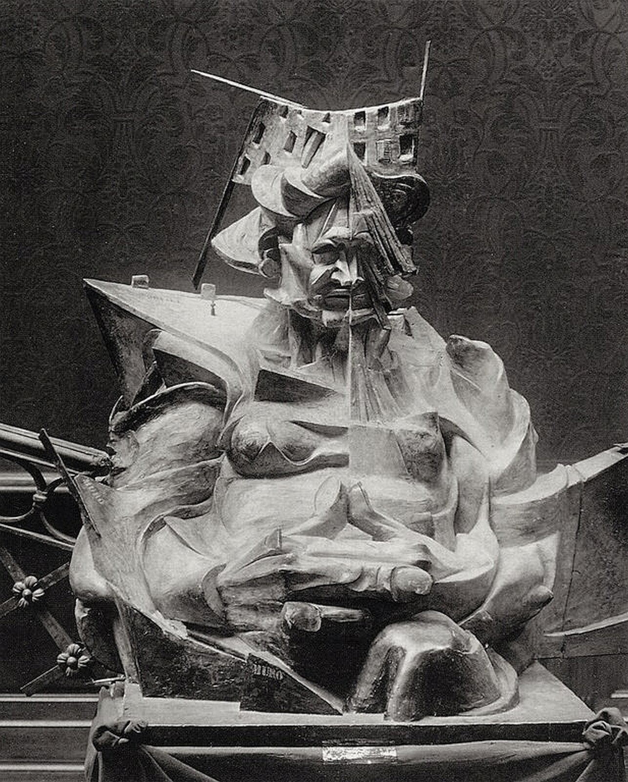 Ilustracja przedstawia rzeźbę Umberta Boccioniego „Głowa + dom + światło”. Ukazuje posąg zdeformowanego człowieka, ujętego od głowy do pasa, połączonego z usytuowanymi pod różnymi kątami płaszczyznami oraz zespolonego z obiektami. Wprowadza to efekty światłocieniowe. Postać na głowie ma dołączony fragment budynku z oknami. Z prawego ramienia wychodzi ostro zakończony fragment powierzchni płaskiej. Abstrakcyjne konstrukcje dołączone są do całej postaci. Mimika człowieka wskazuje na srogość. Z przodu mężczyzna ma zaplecione duże, masywne dłonie.  Rzeźba ustawiona jest na kwadratowej płycie.