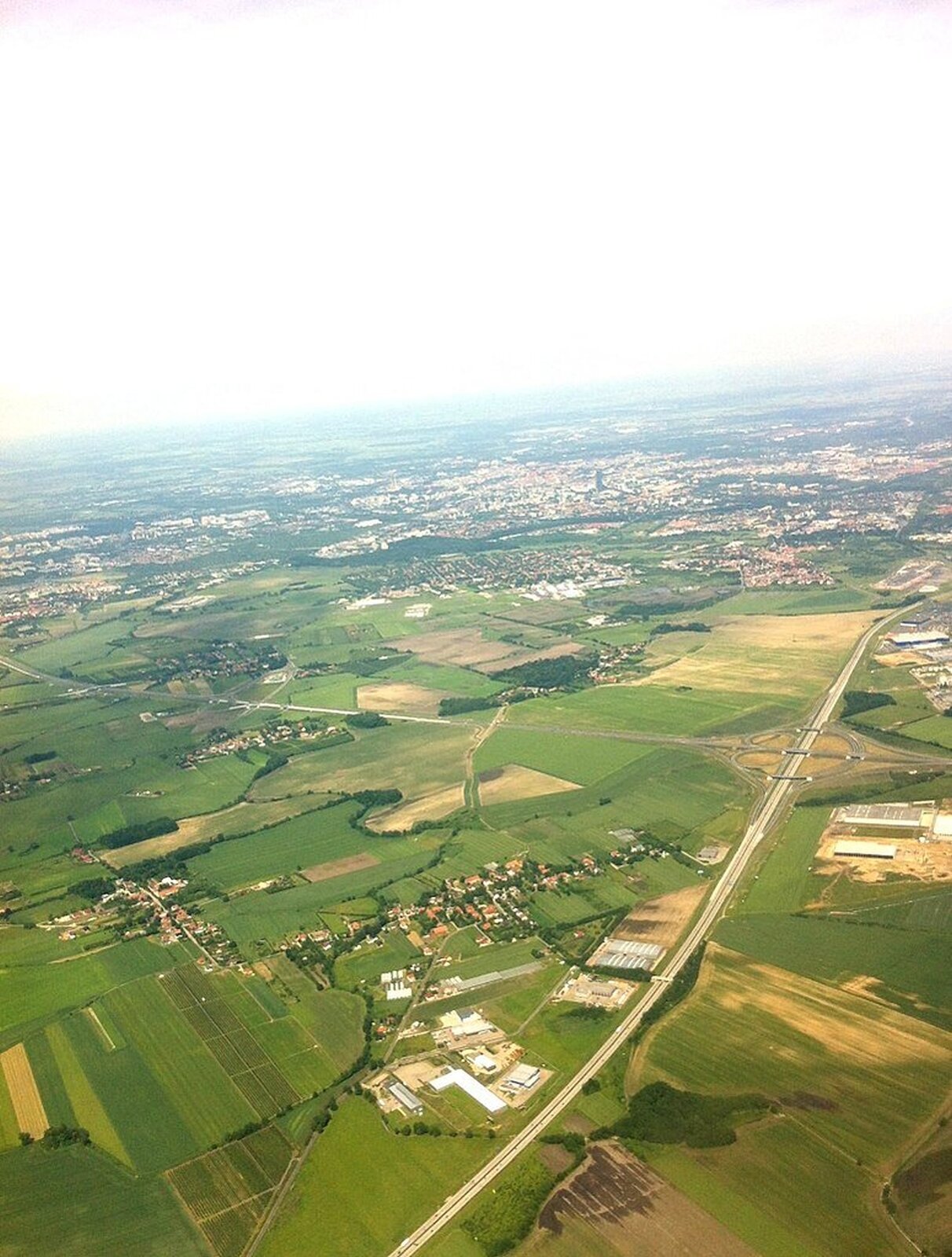 Fotografia lotnicza przedstawiająca na pierwszym planie pola uprawne, między którymi przechodzą dwie krzyżujące się pod kątem prostym autostrady. W miejscu przecięcia się wykonano węzeł pozwalający na zjazd na wybraną drogę. Wzdłuż autostrady gdzieniegdzie budynki i niewielkie osady, na drugim planie miasto.