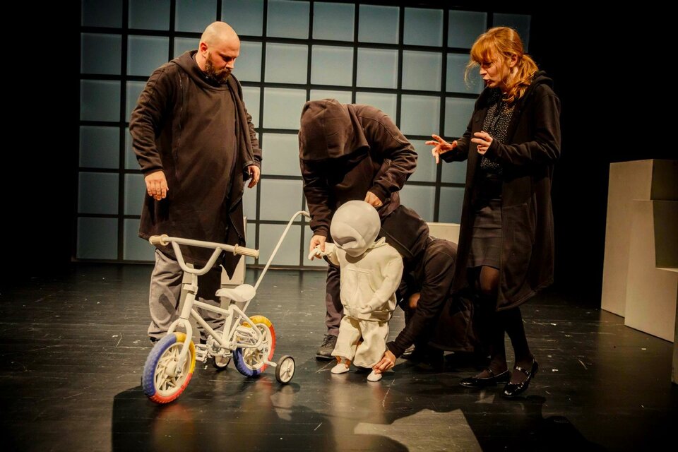 Zdjęcie przedstawia scenę teatralną o minimalistycznej scenografii. Jedynym rekwizytem jest rowerek dziecinny, wyróżnia się też jasna ściana i mała postać, która jest mechaniczną lalką. Czworo aktorów w ciemnych kostiumach przechodzi właśnie test na przyszłych rodziców, opiekując się imitacją dziecka. 