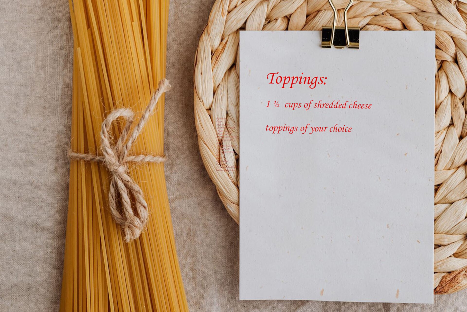 Zdjęcie przedstawia leżące na lnianej, szarej tkaninie: po lewej – makaron spaghetti związany szarym sznurkiem, a po prawej – kartkę przypiętą dużym spinaczem, leżącą na plecionej podkładce. Na kartce widoczny jest przepis na sos w języku angielskim:Toppings: 1 1/2 cups of shredded cheese and toppings of your choice.