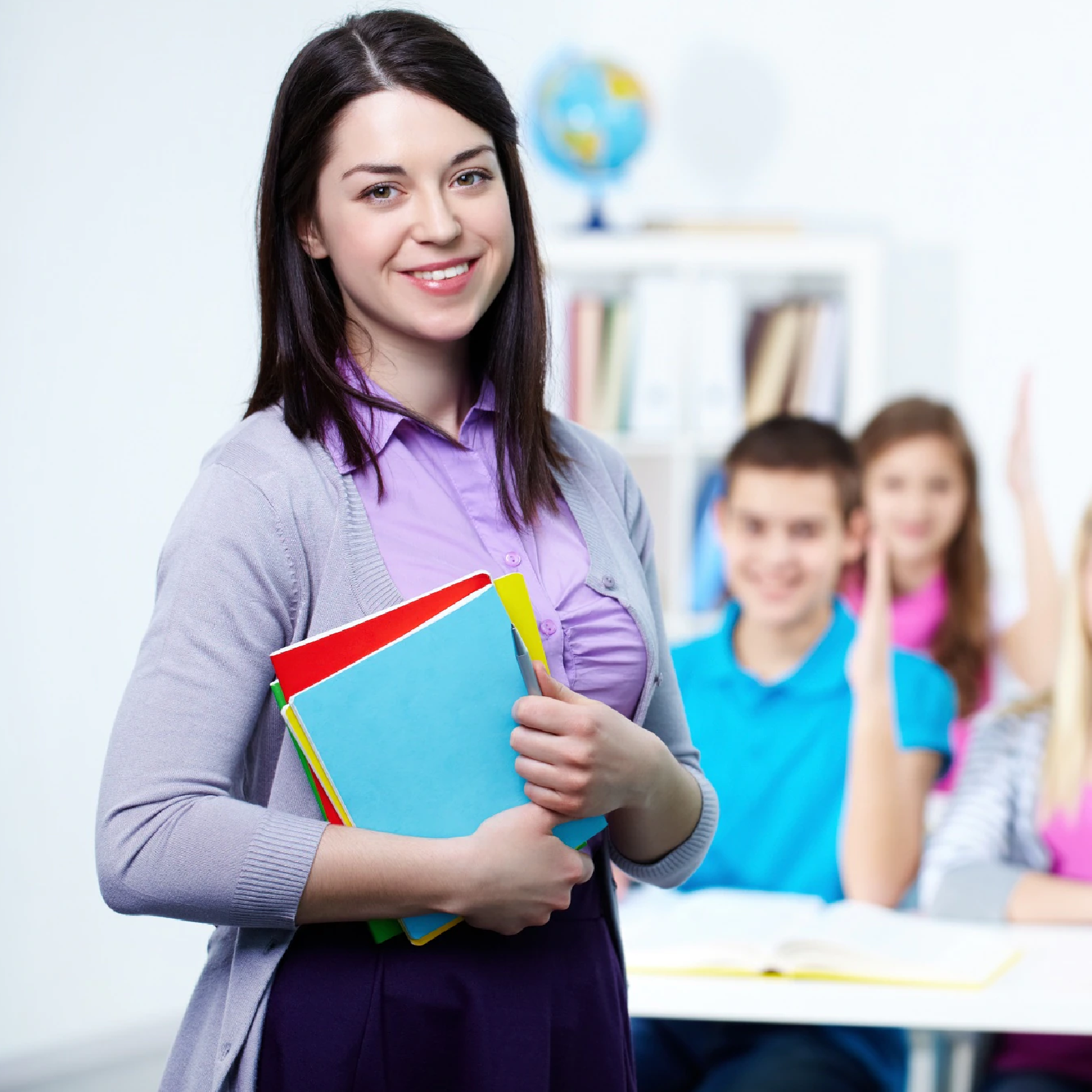 Zdjęcie przedstawia nauczycielkę w klasie, która trzyma w dłoniach kolorowe zeszyty. W tle widoczni są uczniowie w ławkach. 