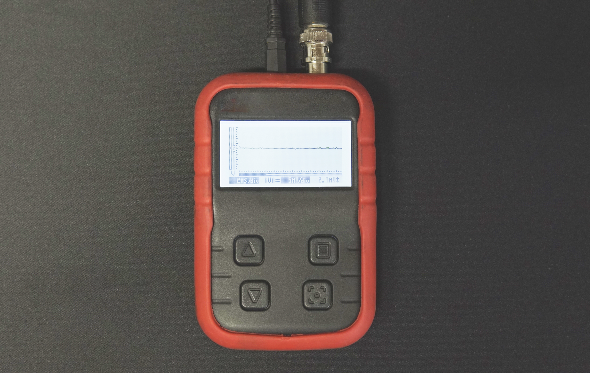 Zdjęcie przedstawia oscyloskop. Urządzenie jest w czerwono‑czarnej obudowie. Posiada wyświetlacz oraz cztery przyciski sterujące. W górnej części są dwa gniazda, do których podłączone są dwa przewody. 