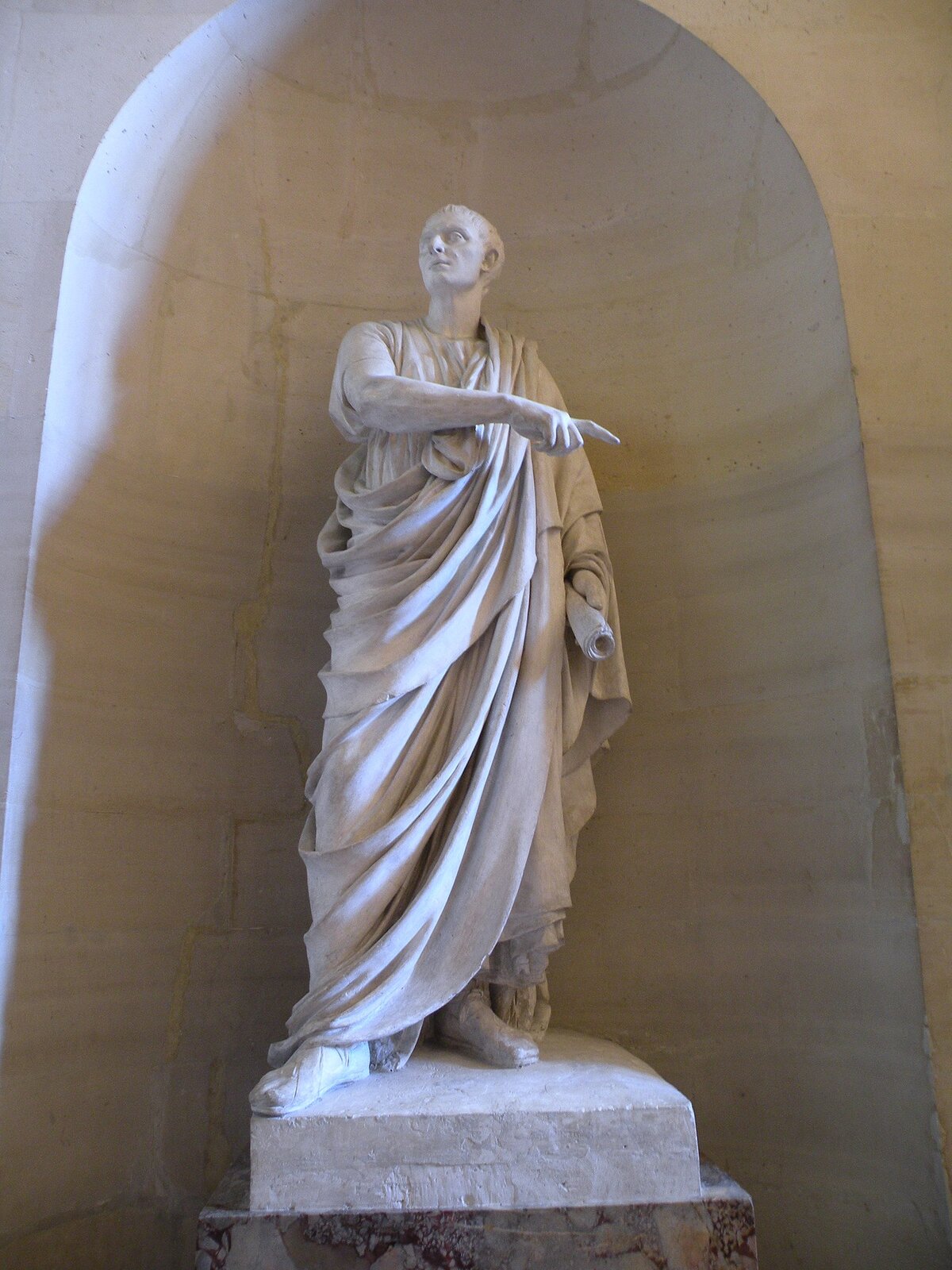 Ilustracja przedstawia rzeźbę ukazującą Cycerona. Rzeźba mężczyzny stoi na kamieniu w zaokrąglonej wnęce w ścianie. Sama postać jest wyprostowana, odziana w długą, sięgającą ziemi szatę. Głowę ma skierowaną w prawą stronę. W lewej ręce trzyma zrolowany papirus, natomiast prawą ręką, palcem wskazującym, wskazuje na coś po swojej lewej stronie.