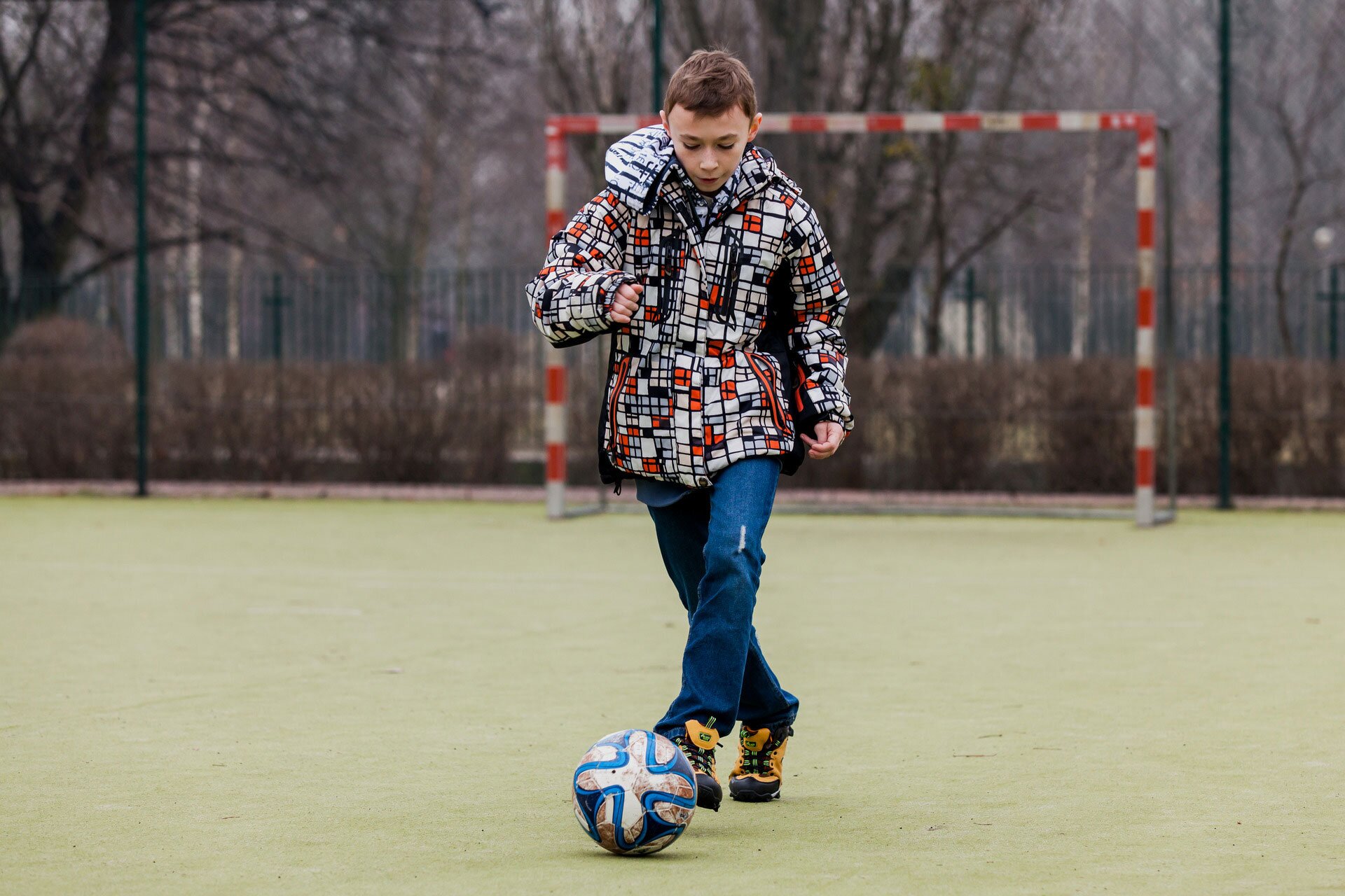 Fotografia przedstawia chłopca na boisku, który kopie piłkę. Za nim stoi bramka. Ubrany jest w kurtkę w czerwono-biało-czarną kratkę, niebieskie dżinsy i żółto-czarne buty. 