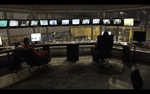 Zdjęcie przedstawia pulpit sterowniczy w kabinie operatorskiej linii zwijania w kręgi. Na pierwszym planie znajduje się dwóch mężczyzn siedzących w fotelach. Obaj mężczyźni ubrani są w pomarańczowe stroje pracownicze. Przed nimi znajduje się biurko z pulpitem sterowniczym. Na biurku znajduje się parę monitorów, wyświetlone są na nich parametry urządzeń. Przy oknach podwieszone są monitory, na który widoczny jest obraz z kamer. Za pulpitem sterowniczym znajduje się ściana złożona z okien. Za oknami widoczna jest hala linii zwijania w kręgi.