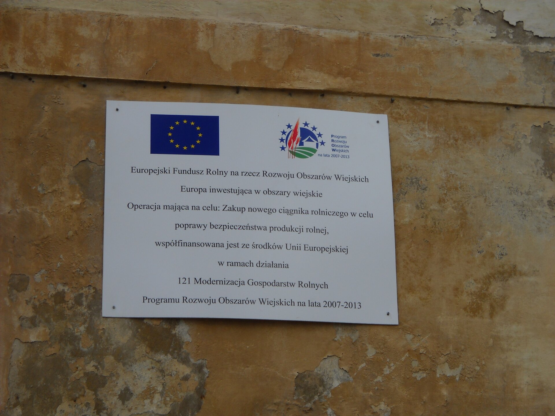 Zdjęcie przedstawia biała tablice wiszące na ścianie budynku. Ściana jest odrapana. Napis na tablicy jest następujący: Europejski Fundusz Rolny na rzecz Rozwoju Obszarów Wiejskich Europa inwestująca w obszary wiejskie. Operacja mająca na celu: zakup nowego ciągnika rolniczego do celu poprawy bezpieczeństwa produkcji rolnej, współfinansowana jest ze środków Unii Europejskiej w ramach działania 121 Modernizacja Gospodarstw Rolnych Programu Rozwoju Obszarów Wiejskich na lata 2007-2013. Na tablicy jest logo unii - na niebieskim prostokącie są żółte gwiazdy tworzące okrąg, a także logo Programu Rozwoju Obszarów Wiejskich - logo jest okrągłe, jest na nim dom, fragment pola, trzy trawy. Górną część logo otaczają niebieskie gwiazdki.