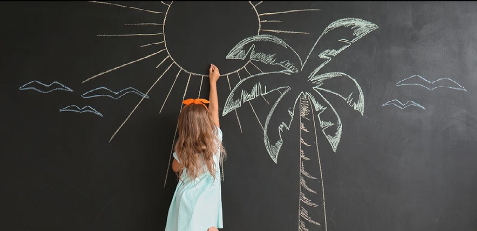 Zdjęcie przedstawia małą dziewczynkę o długich włosach, związanych pomarańczową tasiemką. Nosi bladoniebieską sukienkę z krótkimi rękawami. Stoi przodem do szkolnej tablicy i prawą ręką rysuje wielkie słońce z promykami, palmę i pięć lecących ptaków. Rysunek jest bardzo duży.