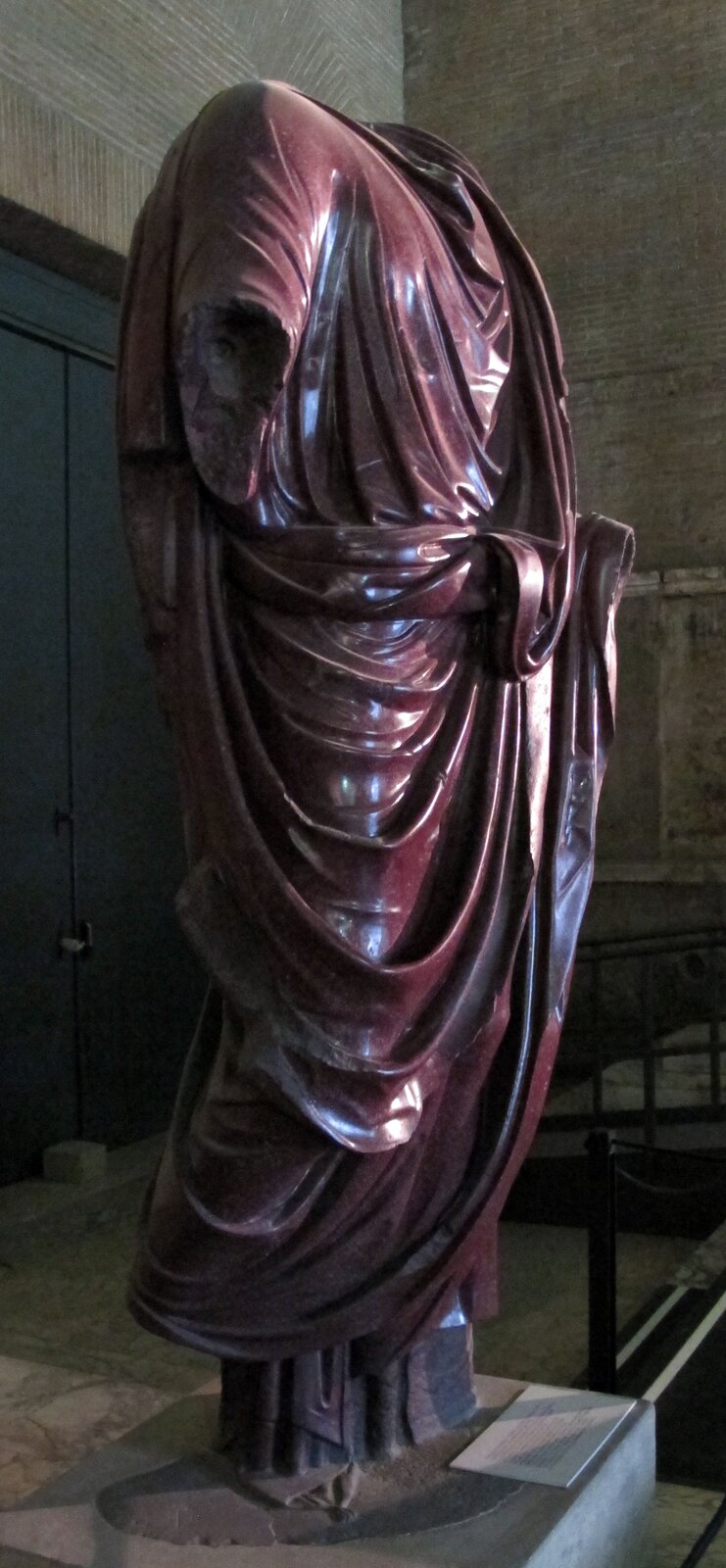 Ilustracja przedstawia rzeźbę mężczyzny ubranego w długą szatę. Posąg jest uszkodzony: brak jest głowy i rąk. Szata jest precyzyjnie udrapowana. Na uwagę zasługuje jej purpurowy kolor przysługujący jedynie dostojnikom państwowym.