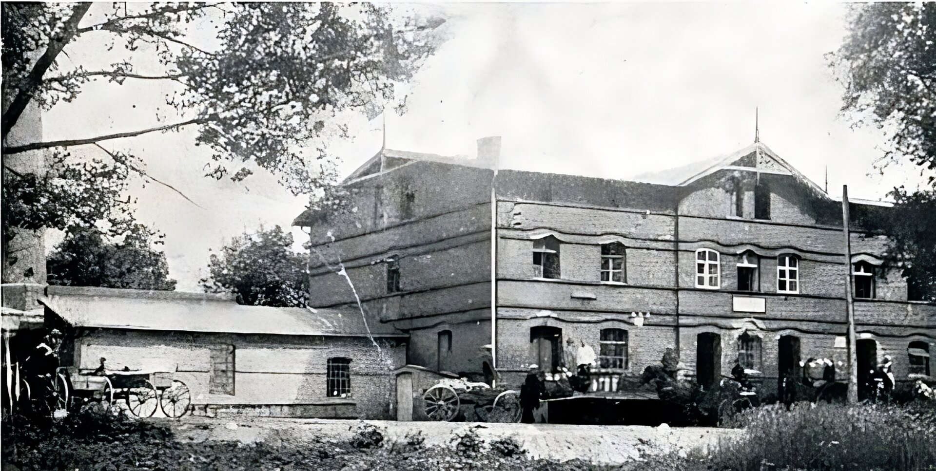 Czarno‑białe zdjęcie. Ceglany, dwupiętrowy budynek. z niższą przybudówką po lewej stronie.  Przed budynkiem stoją ludzie oraz powozy. 