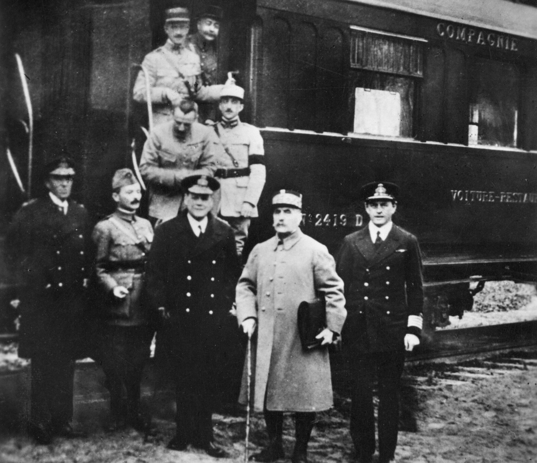 Zdjęcie przedstawia żołnierzy na tle wagonu pociągu. Przed stoi pięciu mężczyzn, kolejni czterej stoją za nimi, na schodach i wysiadając na peron. Wszyscy ubrani są w mundury wojskowe. Pierwszy, trzeci i piąty od prawej mają ciemne dwurzędowe marynarki od mundurów, ciemne spodnie, czapki z daszkiem, białe koszule i ciemne krawaty. Starszy mężczyzna z wąsami w pierwszym rzędzie (drugi od prawej) stoi wsparty o laskę, w drugim ręku trzyma teczkę. Ma na sobie jasny wojskowy płaszcz, na głowie czapkę z daszkiem. Wszyscy pozostali żołnierze także ubrani są w jasne garnitury.