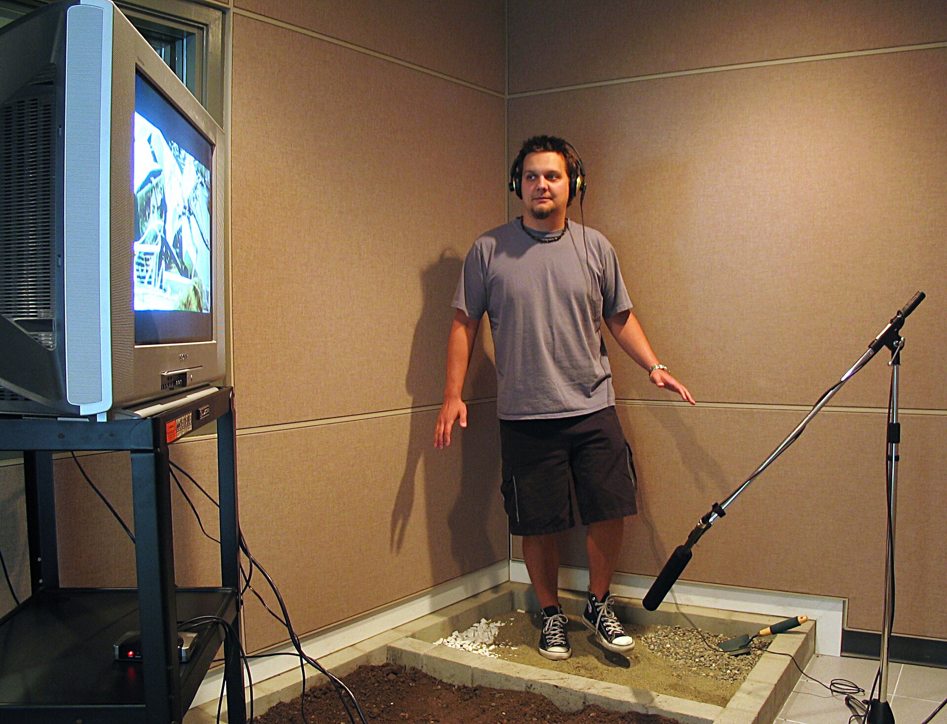 Zdjęcie przedstawia mężczyznę podczas nagrywania efektów synchronicznych w studio. Stoi on w w rogu sali w wydzielonej przestrzeni o powierzchni około jednego metra kwadratowego wypełnionej piaskiem. Mężczyzna ma na sobie słuchawki nauszne, ubrany jest w koszulkę z krótkim rękawem, krótkie spodenki i trampki. Po lewej stronie w kadrze pod ostrym kątem znajduje się ekran wyświetlający nagranie. Po prawej stronie na statywie zamontowany jest mikrofon.