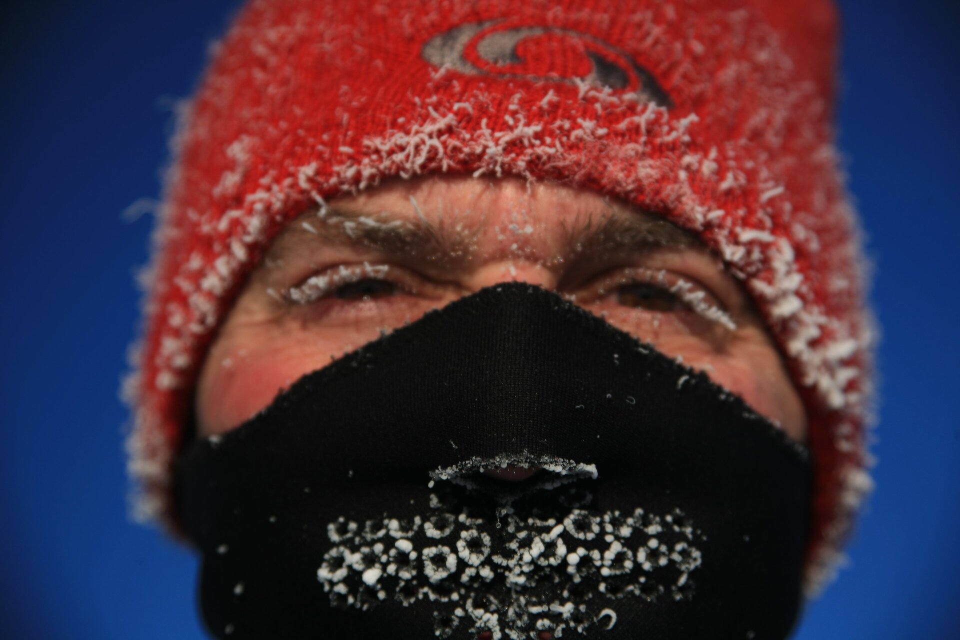 Zdjęcie przedstawia twarz mężczyzny. Na głowie ma wełnianą czapkę. Usta oraz nos zakrywa wełnianym szalem. Widać tylko oczy i krzaczaste brwi. Na rzęsach, brwiach oraz na czapce w jej dolnej części osadził się szron.     