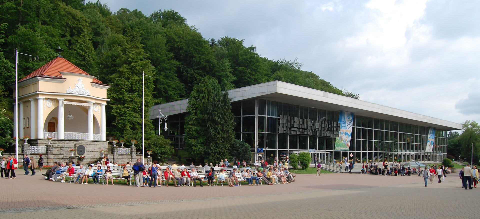 Na zdjęciu oszklony podłużny budynek z płaskim dachem. Przed budynkiem rozległy plac wyłożony kostką. Rozmieszczone są ławki, siedzi na nich dużo osób, pozostali przechadzają się. Za budynkiem linia drzew liściastych.