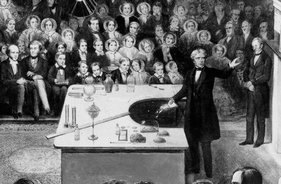 Rys. 3. Na pierwszym planie ilustracji znajduje się duży stół, zastawiony wieloma przyrządami do demonstracji zjawisk elektrycznych. Za stołem stoi Faraday, który wskazuje ręką na tablicę znajdująca się za nim. Z tyłu w wielu rzędach siedzi publiczność. Kobiety i mężczyźni ubrani są według XIX‑wiecznej mody.