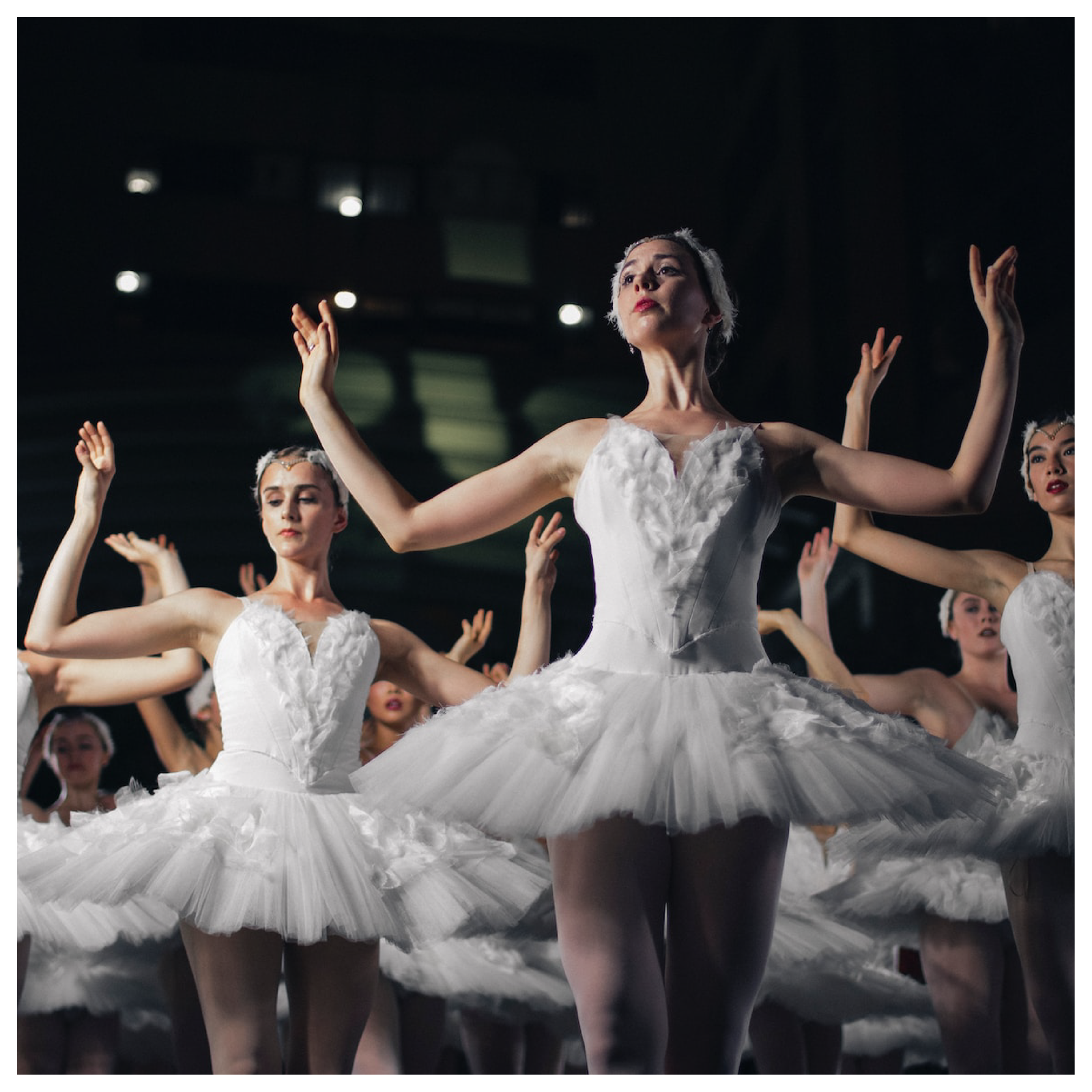 Ilustracja przedstawia występ baletowy. Na scenie grupa kobiet - baletnic, w takich samych, białych strojach. Wszystkie w tej samej pozycji z dłońmi uniesionymi po bokach na wysokości głowy.