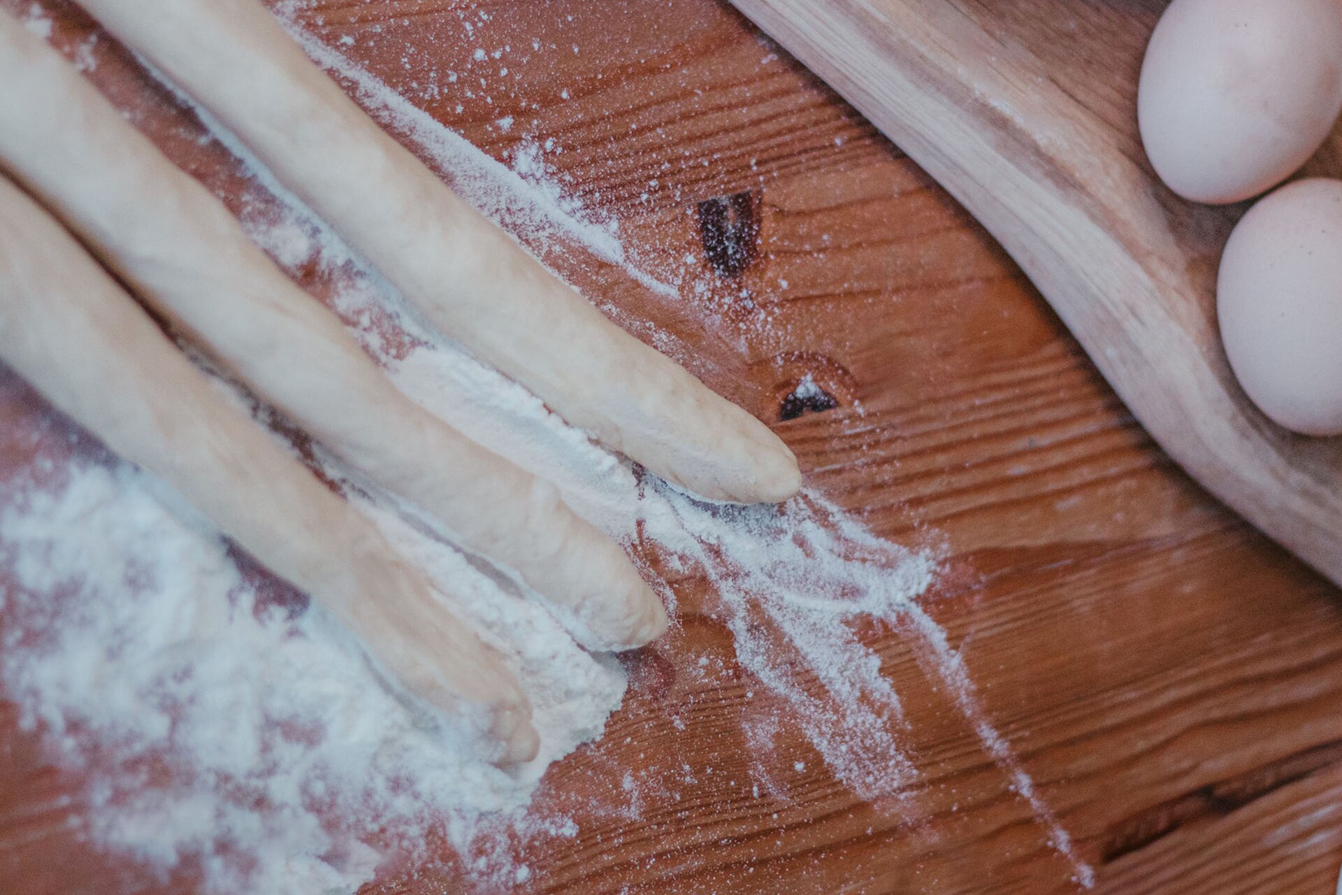 Zdjęcie przedstawia krok drugi i trzeci formowania chały tureckiej. Zdjęcie wykonane jest od góry pod skosem. Widoczny jest ciemnobrązowy drewniany blat na którym w lewej części grafiki znajdują się trzy owalne podłużne wałki ciasta oprószone białą mąką. W prawym górnym rogu zdjęcia dostrzec można jasnobrązową drewnianą deskę ułożoną pod skosem. Na niej widoczne są dwa jajka.