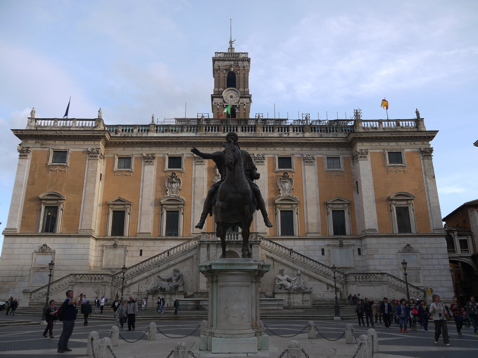 Zdjęcie przedstawia budynek. Posiada dwie kondygnacje, prostą bryłę, płaski dach, prostokątne okna, attykę, wieżę z zegarem. Ma wysoką podmurówkę, do której przylega balkon. Do budynku prowadzą schody znajdujące się po dwóch stronach balkonu. Budynek stoi na wzgórzu. Przed budynkiem chodzą ludzie. Na placu przed budynkiem, na piedestale, stoi pomnik mężczyzny siedzącego na koniu z wyciągniętą prawą dłonią przez siebie.