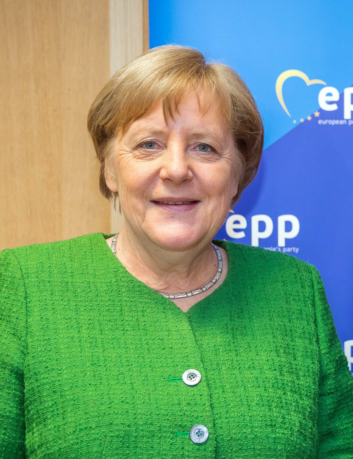 Zdjęcie przedstawia Angelę Merkel. To dojrzała, jasnowłosa kobieta. Ma owalną twarz, na której wyraźnie zaznaczone są zmarszczki. Kobieta ma krótkie, proste włosy. Nieznacznie się uśmiecha. Ubrana jest w żakiet.