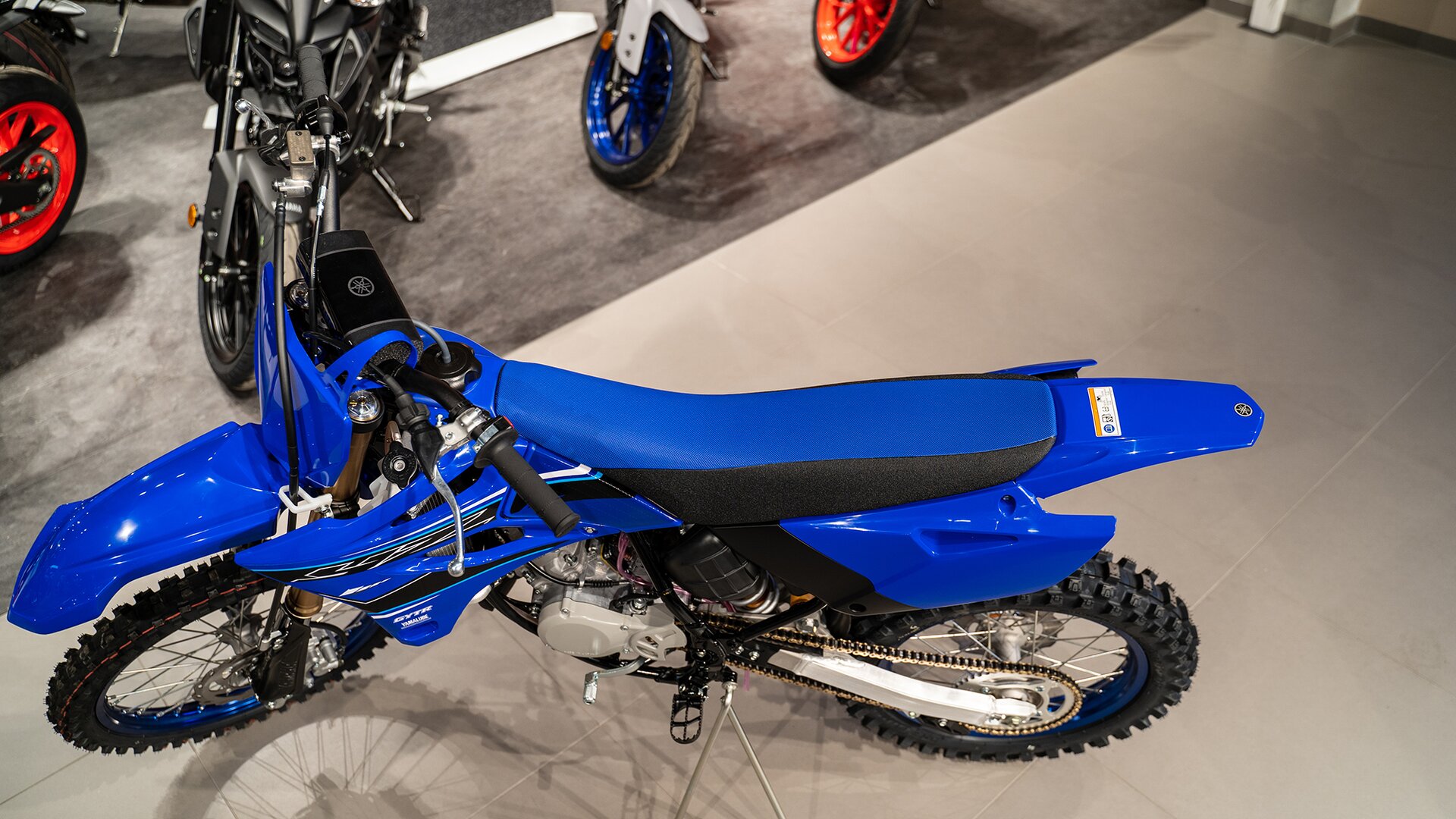 Zdjęcie przedstawia motocykl - widok z góry - o lekkiej ramie. Motocykl jest wąski, ma bardzo wąskie siedzenie, wysoko nad kołami uniesione błotniki. Bieżnik na oponach jest głęboki. Pojazd ma niebieską ramę.  