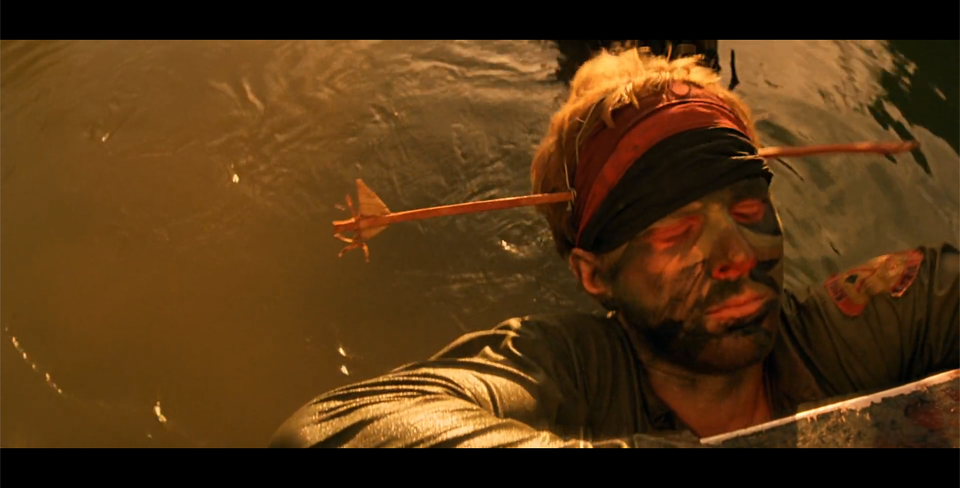 Kadr przedstawia mężczyznę, który jest zanurzony w wodzie. Ukazana jest jego głowa. Mężczyzna ma zamknięte oczy. Ma założoną na głowę strzałę, która wydaje się, jakby przebiła mu głowę na wylot. Mężczyzna ma na twarzy namalowane ciemnozielone plamy.