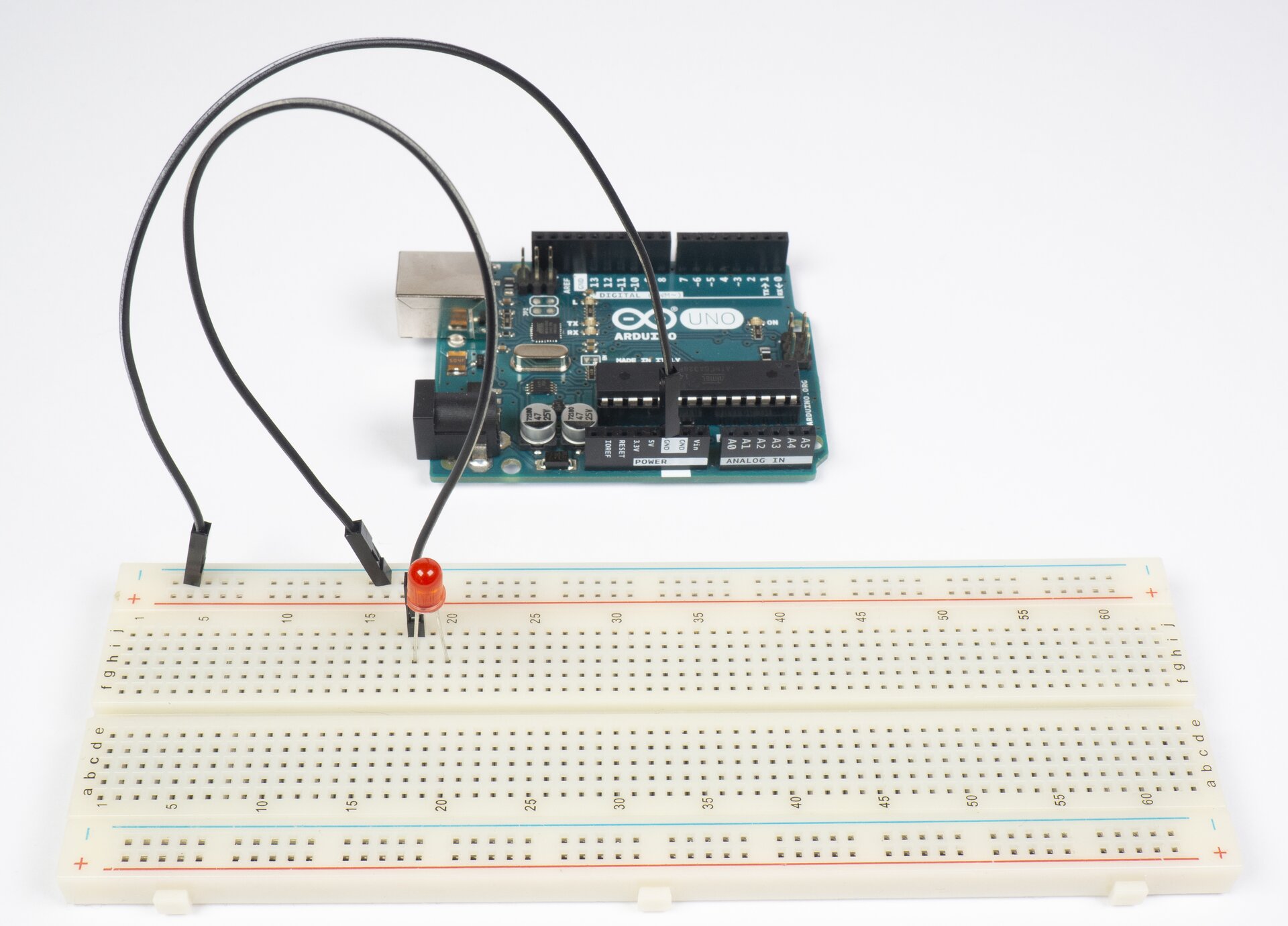 Zdjęcie przedstawia płytkę Arduino oraz sześćdziesięciowierszową płytkę stykową. Wiersze płytki stykowej podpisano co pięć zaczynając od 1. Po prawej i po lewej stronie podpisano poszczególne kolumny. Na górnym obszarze: j i h g f. Na dolnym obszarze: e d c b a. Na płytce stykowej znajduje się czerwona dioda LED oraz czarny przewód. Ze złącza GND Arduino wyprowadzono czarny przewód, którego koniec umieszczono w drugim otworze szyny z oznaczeniem minus. Jeden koniec drugiego czarnego przewodu umieszczono w otworze na szynie minus, a drugi koniec umieszczono w kolumnie j w wierszu 18. Jedną nogę diody LED umieszczono w kolumnie h w wierszu 18, a drugą nogę w kolumnie h w wierszu 20.