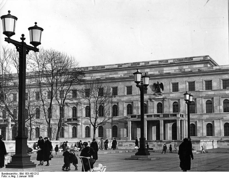Na zdjęciu jest duży dwupiętrowy budynek. Nad wejściem jest wizerunek orła. Przy budynku jest plac, po którym spacerują ludzie.