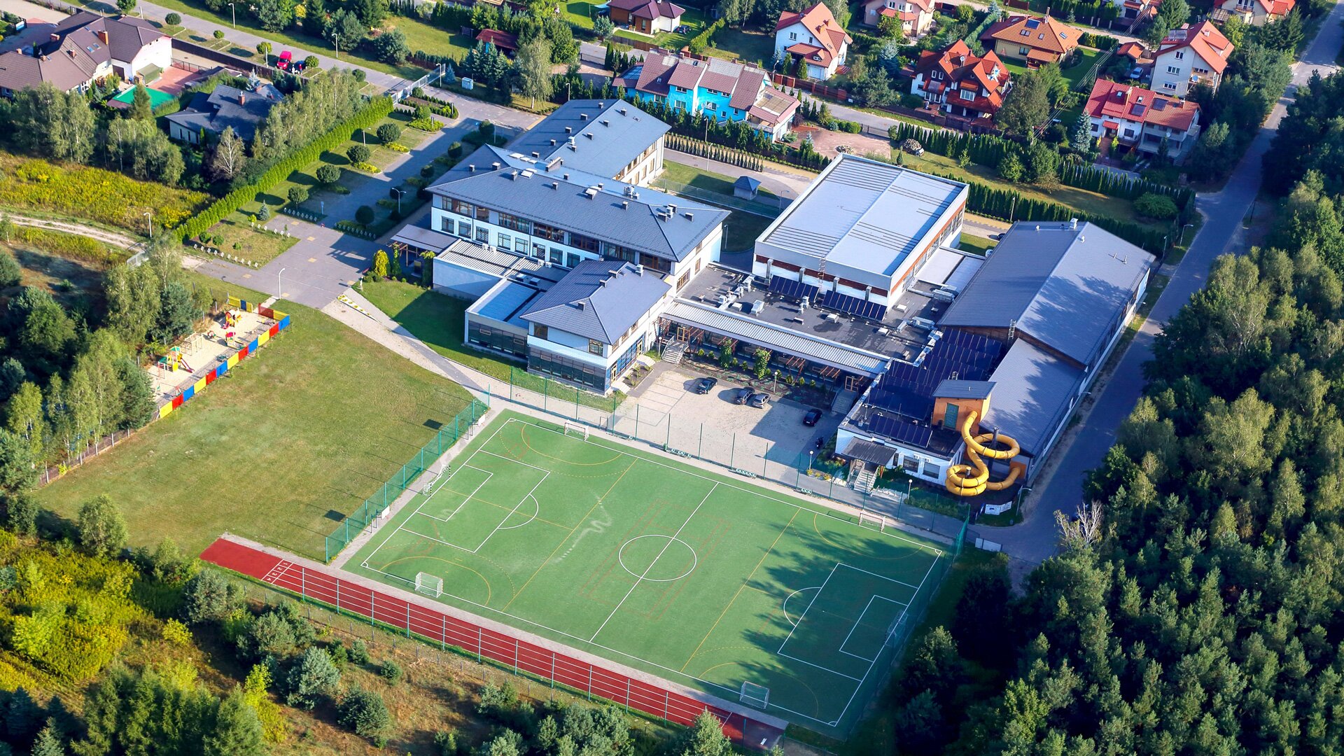 Zdjęcie przedstawia widok z góry na budynek. Obok niego jest boisko piłkarskie, bieżnia oraz domy jednorodzinne. Widoczne są ulice prowadzące do szkoły i domów jednorodzinnych oraz fragment lasu. 
