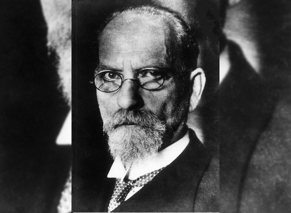 Zdjęcie przedstawia starszego mężczyznę. Jest łysiejący, ma pociągłą twarz i nosi okulary w okrągłych oprawkach. Ma brodę z wąsami. Ubrany jest w garnitur.