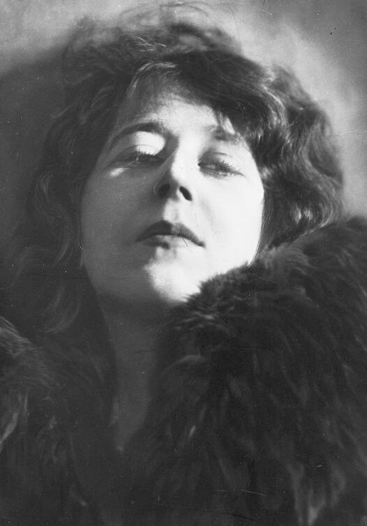 Fotografia przedstawiająca kobietę w sile wieku w ujęciu od piersi w górę. Kobieta ma owalną twarz. Unosi głowę. Jej brwi są ciemne. Oczy lekko przymknięte. Postać spogląda w dół. Ma duży, prosty nos. Usta kobiety są wydatne. Poetka unosi podbródek. Ma ciemne, gęste włosy sięgające ramion. Jest ubrana w futro.