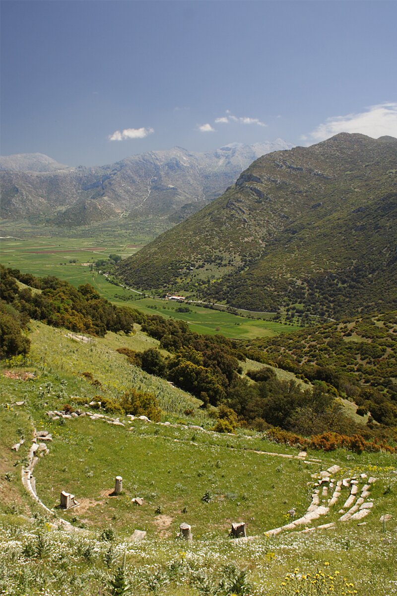 Zdjęcie przedstawia krajobraz górski. Widok na porośniętą zieloną roślinnością dolinę i niedalekie zielone wzniesienia, na horyzoncie wysokie pasmo górskie. W dole droga. Z przodu pozostałości kamiennej zabudowy w kształcie półokręgu. 