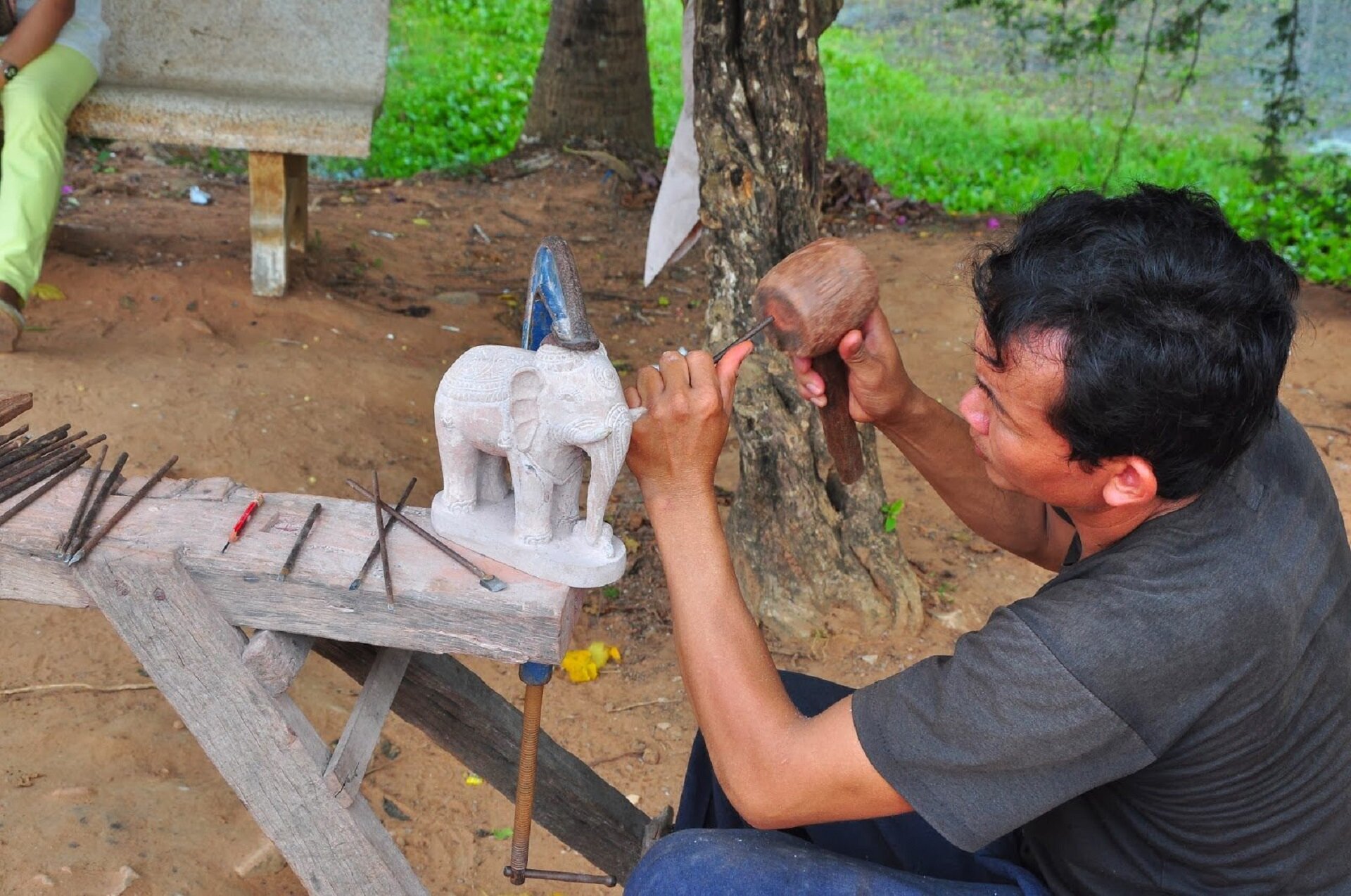 Zdjęcie przedstawia mężczyznę, który rzeźbi w z kamienia. Rzeźba ukazuje słonia w kolorze szarym.