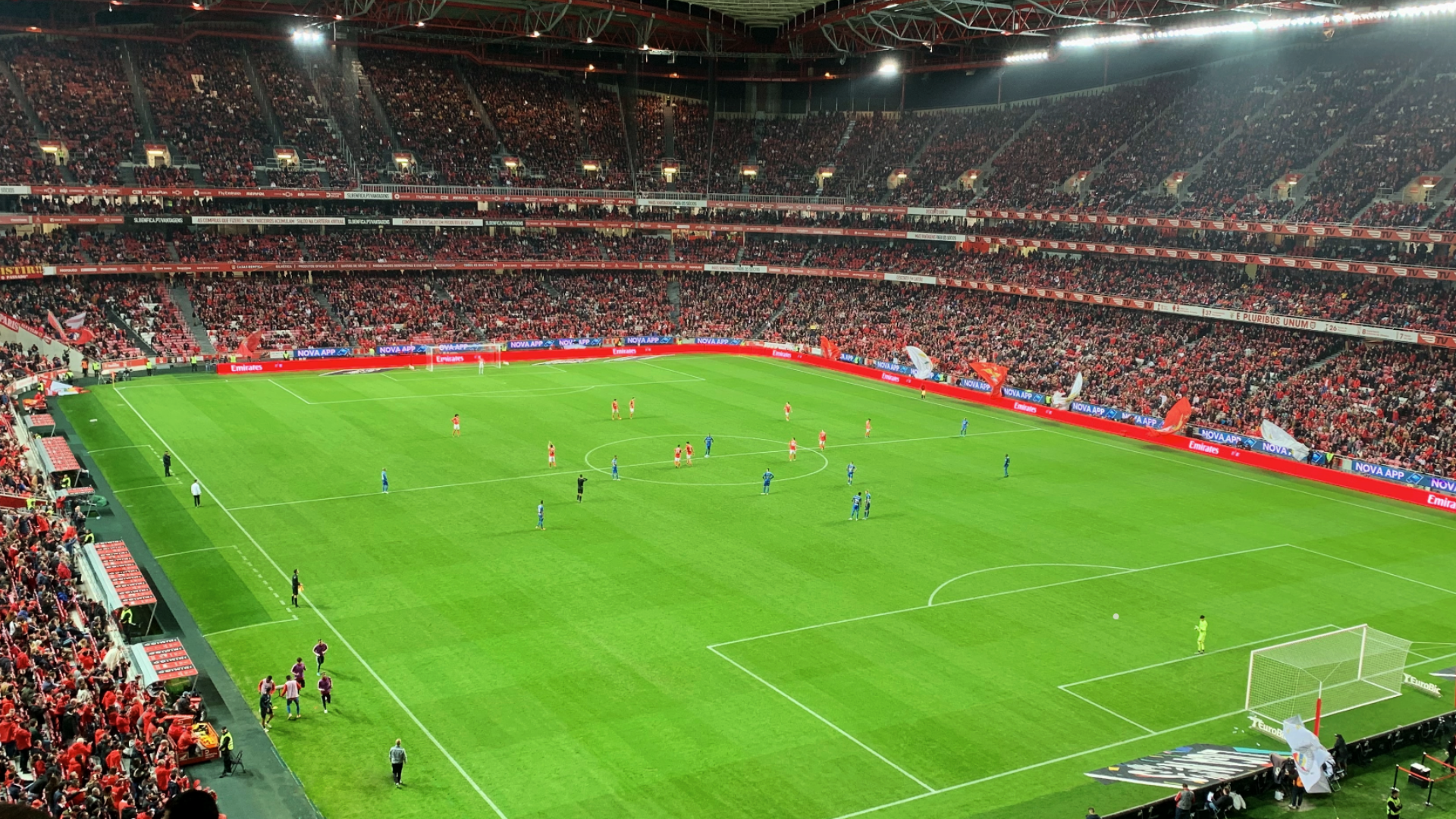 Zdjęcie przedstawia oświetlony, pełen kibiców stadion w czasie rozgrywania meczu piłki nożnej.