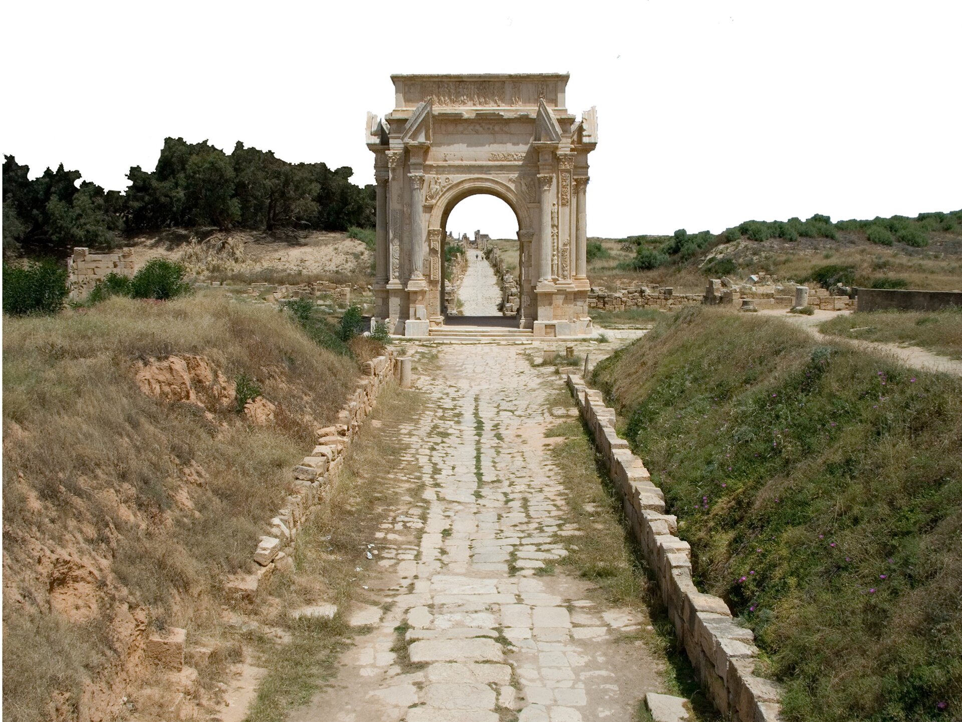 Na ilustracji fragment wybrukowanej drogi, zakończonej łukiem triumfalnym. Po obu stronach drogi nasyp ziemny, ten z prawej porośnięty trawą. W tle drzewa.