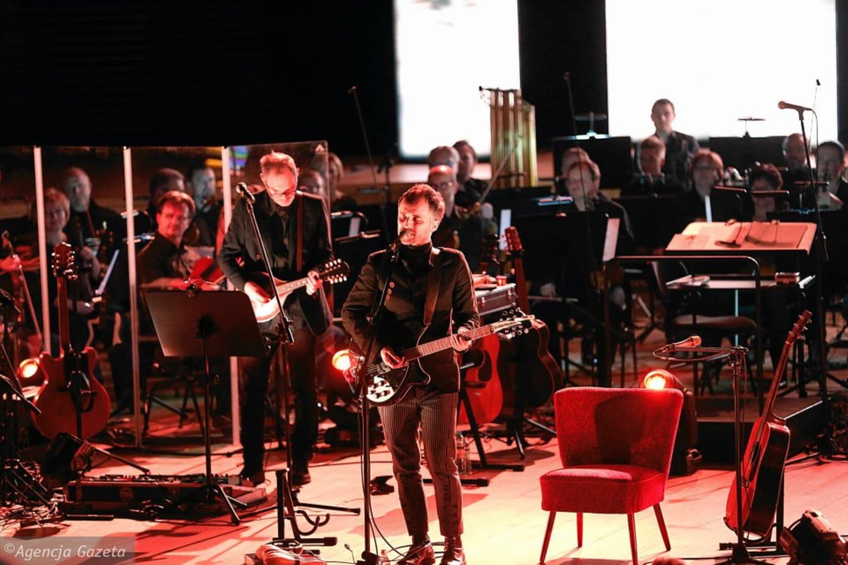 Artur Rojek wraz z Narodową Orkiestrą Symfoniczną Polskiego Radia Fotografia przedstawia scenę. Na pierwszym planie uchwycono mężczyznę ubranego na czarno, grającego na czarno‑białej gitarze elektrycznej. Przed nim stoi statyw wraz z mikrofonem. Za nim jest około piętnastu muzyków, z których większa część siedzi przed instrumentami i pulpitami z nutami. Z przodu, po prawej stronie, stoi niski fotel bez podłokietników, a z lewej – instrumenty muzyczne. Na scenie są trzy lampy, oświetlające ją ciepłym światłem.