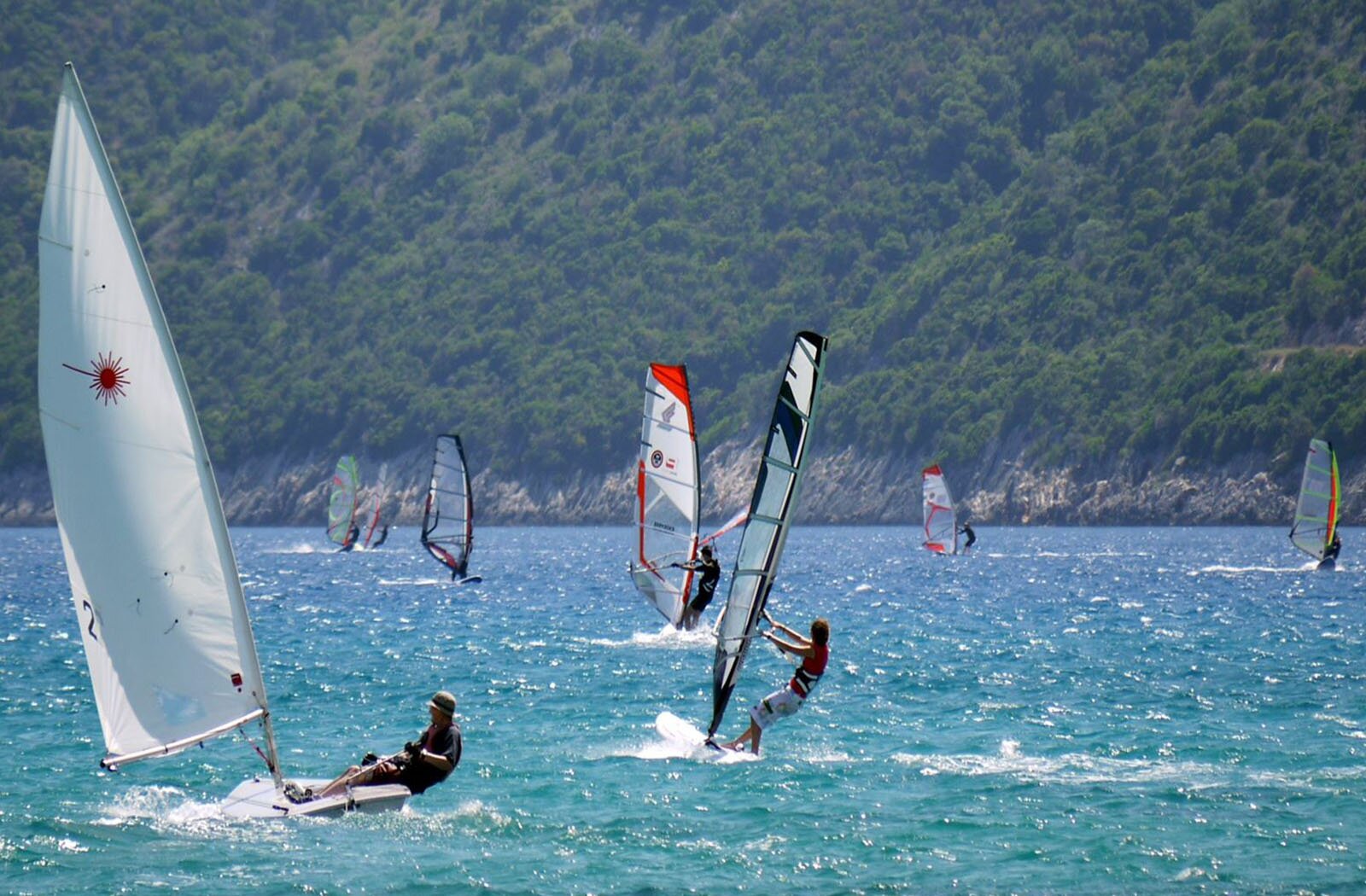 Zdjęcie przedstawia zbiornik wodny otoczony skalistymi górami porośniętymi zielonymi drzewami. Na akwenie widać osiem osób, które uprawiają windsurfing. Widoczne są żagle w różnych kolorach.