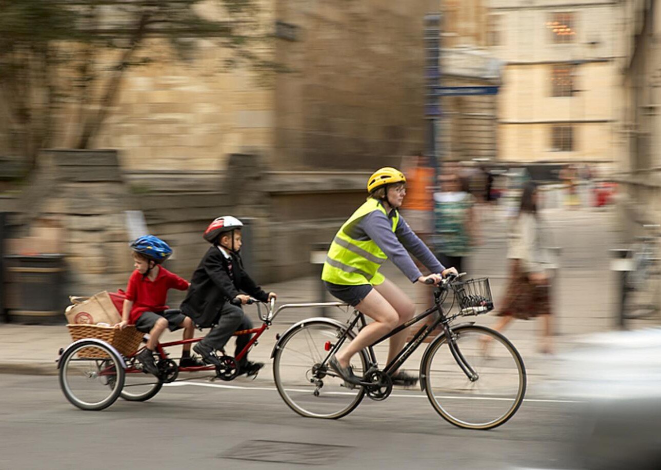 Fotografia barwna. Kobieta w kamizelce odblaskowej i kasku na głowie jedzie rowerem, do którego jest przyczepiona przyczepka imitująca rower typu tandem dla dzieci. Z tyłu siedzi dwóch chłopców w kaskach na głowach. Za ostatnim siodełkiem znajduje się kosz z zakupami. Tło zdjęcia jest rozmazane, ale na widać, że rowerzyści poruszają się po ulicy miasta.