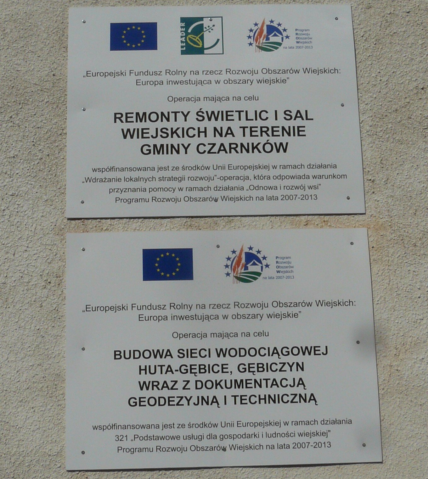 Zdjęcie przedstawia przymocowane do ściany budynku dwie białe tablice z unijnymi symbolami. Tablice informują o operacjach w ramach Europejskiego Funduszu Rolnego na rzecz Rozwoju Obszarów Wiejskich: Europa Inwestująca w obszary wiejskie. Jedna z tablic dotyczy operacji mającej na celu remont świetlic i sal wiejskich na terenie gminy Czarnków, a druga budowy sieci wodociągowej Huta-Głębice, Gębiczyn wraz z dokumentacją geodezyjną i techniczną.
