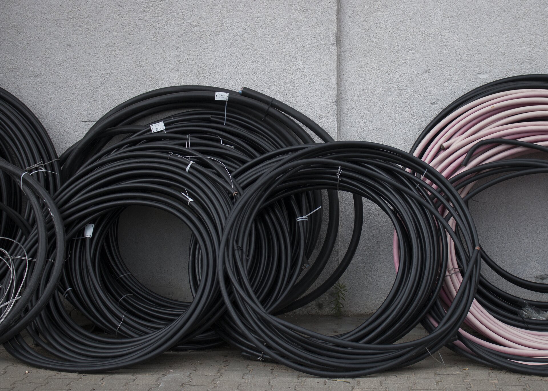 Ilustracja przedstawia zwinięte gumowe węże w kolorze czarnym i jasnoróżowym, oparte o szarą fasadę budynku. Kable ustawione są na chodniku, zbudowanym z szarej kostki.