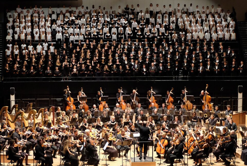Orkiestra symfoniczna i duży chór występują na scenie koncertowej. Dyrygent stoi tyłem do publiczności, przed muzykami grającymi na instrumentach smyczkowych, dętych, harfach i kontrabasach. Chórzyści stoją na podwyższeniu w tle, ubrani głównie na biało i czarno.