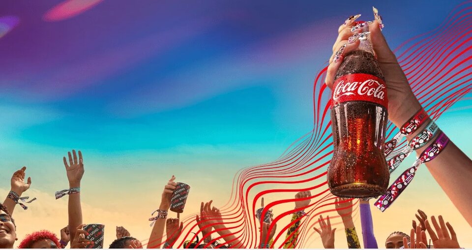 Dłoń trzyma szklaną butelkę Coca‑Cola nad tłumem ludzi z podniesionymi rękami i festiwalowymi opaskami; w tle widać kolorowe niebo i graficzne czerwone linie wychodzące od butelki, tworzące dynamiczny, reklamowy efekt.