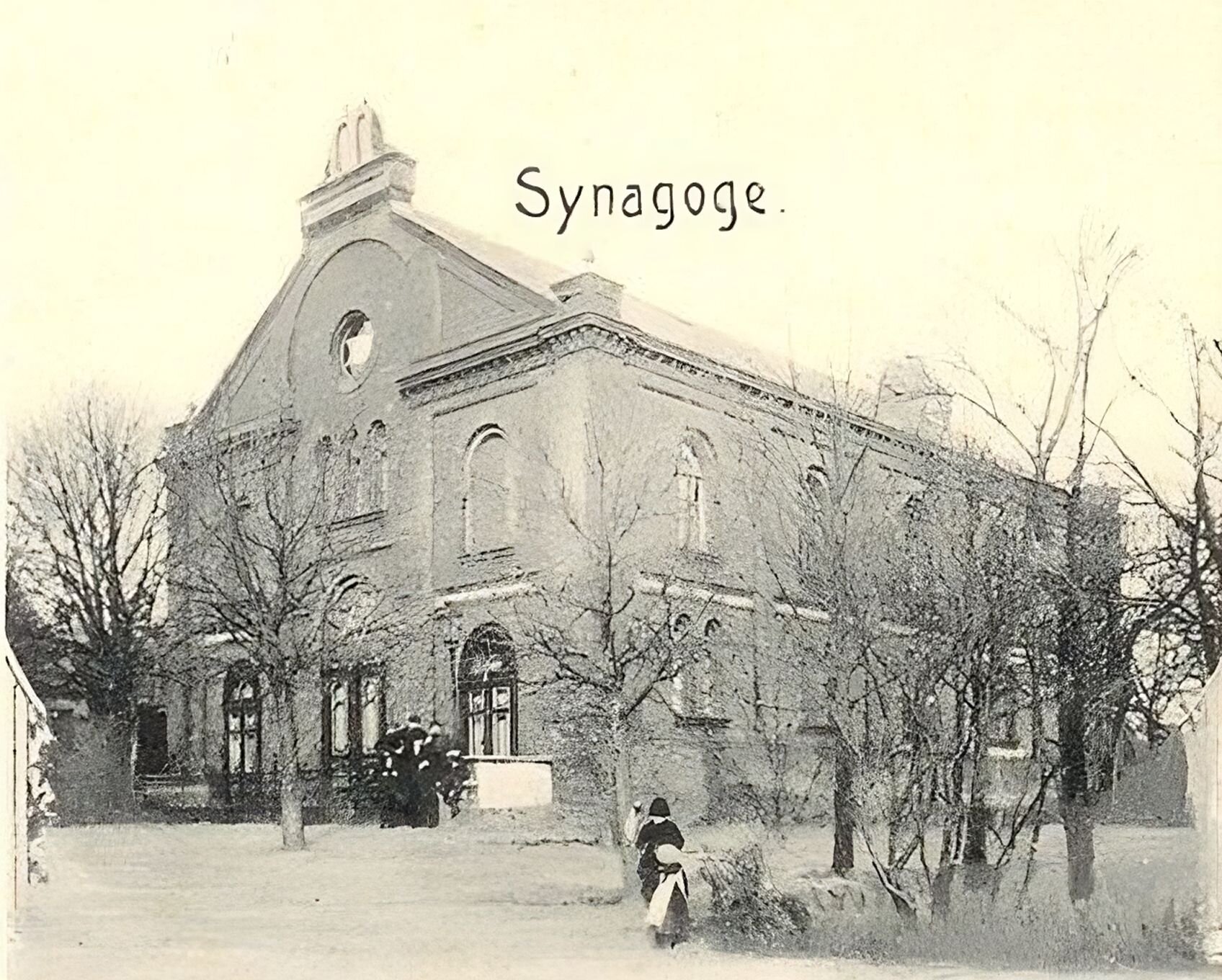 Kliknij, aby powiększyć. Zdjęcie przedstawia budynek o prostej, prostokątnej bryle ze spadzistym dachem. Posiada jedno piętro. Okna w ścianach są duże i zakończone łukiem. Widoczne są dwie ściany – jedna z nich jest frontowa. W centralnej części ma dwuskrzydłowe drzwi, a z obu jego stron mniejsze, również dwudrzwiowe. Na piętrze widoczne są otwory wyglądające jak okna, a nad nimi okrągłe okno. Przed drzwiami stoi kilku ludzi. Wokół budynku rosną bezlistne drzewa.