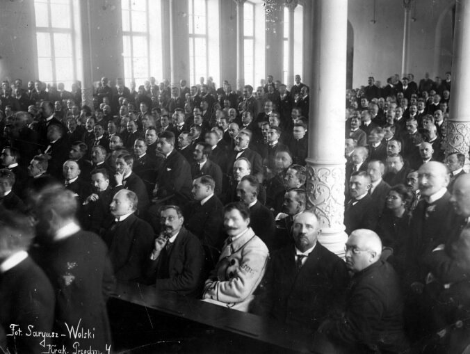 Na zdjęciu w wysokiej sali siedzą przy ławach mężczyźni w garniturach. Ławy są ustawione równolegle wzdłuż sali.