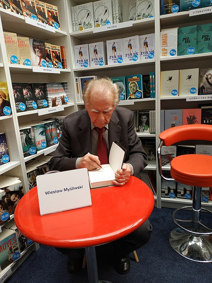 Zdjęcie przedstawia starszego, siwowłosego mężczyznę siedzącego przy niewielkim, okrągłym stoliku. Mężczyzna zapisuje coś na stronie tytułowej książki. Na stoliku stoi kartka z napisem Wiesław Myśliwski. W tle regały wypełnione  książkami.