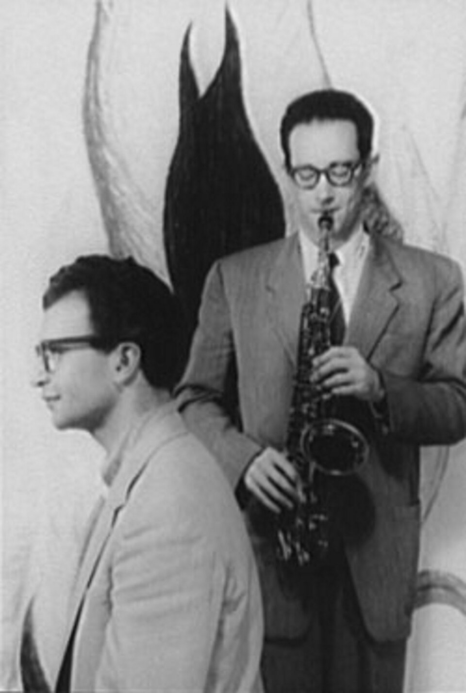 Ilustracja przedstawia dwóch mężczyzn w okularach - Paul Desmond i Dave Brubeck. Jeden mężczyzna siedzi profilem do kadru zdjęcia. Drugi mężczyzna stoi i gra na trąbce. Ubrani są w szare garnitury.