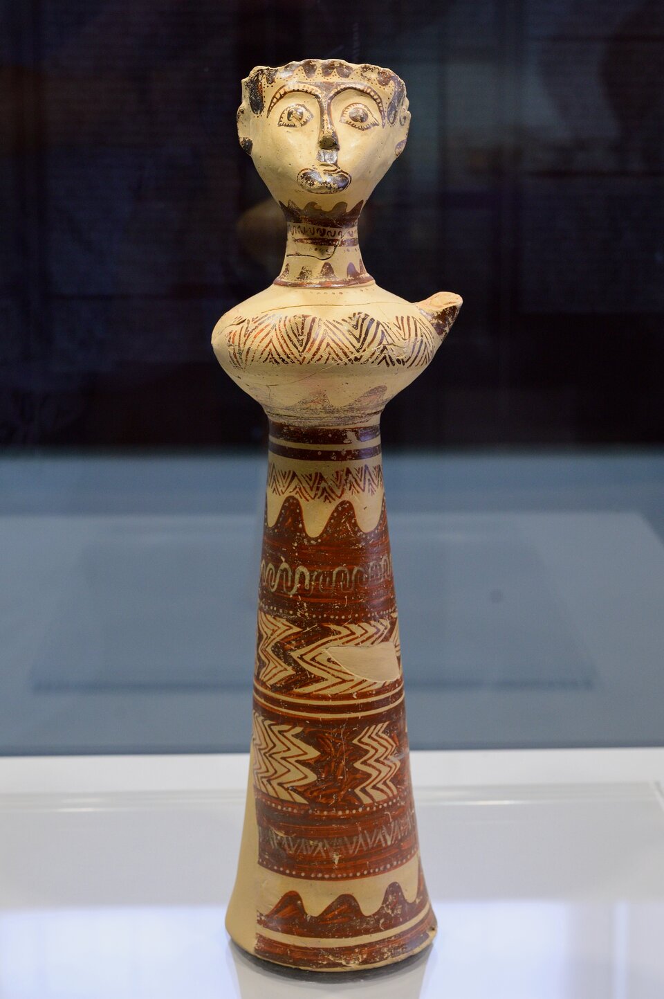 Fotografia przedstawia figurkę "Pani z Phylakopi". Ukazuje kobietę stojącą w wyprostowanej, frontalnej pozie, z ledwie zarysowanymi ramionami uniesionymi do góry wskazującymi na w gest modlitewny lub rytualny. Ubrana jest w charakterystyczny dla epoki bogato zdobiony strój: długą suknię o poziomych pasach. Jej głowa jest proporcjonalnie niewielka, z wyraziście zaznaczonymi rysami twarzy - dużymi, otwartymi oczyma, mocno zarysowanymi brwiami i nosem oraz symboliczną koroną. 