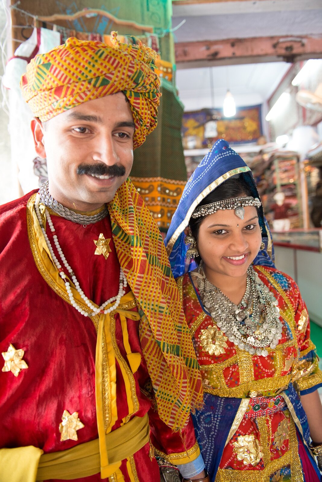 Zdjęcie przedstawia małżeństwo pochodzące z Indii. Ubrani są w ludowe stroje. Mężczyzna ma na głowie turban, kobieta chustę oraz opaskę na głowie z biżuterii i wiele naszyjników. Oboje mają bardzo kolorowe stroje. 